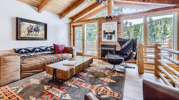 Interior acogedor de una cabaña de madera con un sofá de cuero, una mesa de centro de madera, un sillón de cuero negro y un ventanal con vistas al bosque.