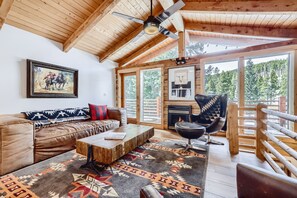 A cozy log cabin interior with a leather sofa, a wooden coffee table, a black leather chair, and a large window offering a forest view.