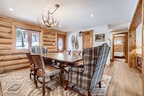 Een chaletinterieur met een eettafel, stoelen, een gestreept fauteuil, houten wanden en een kroonluchter.