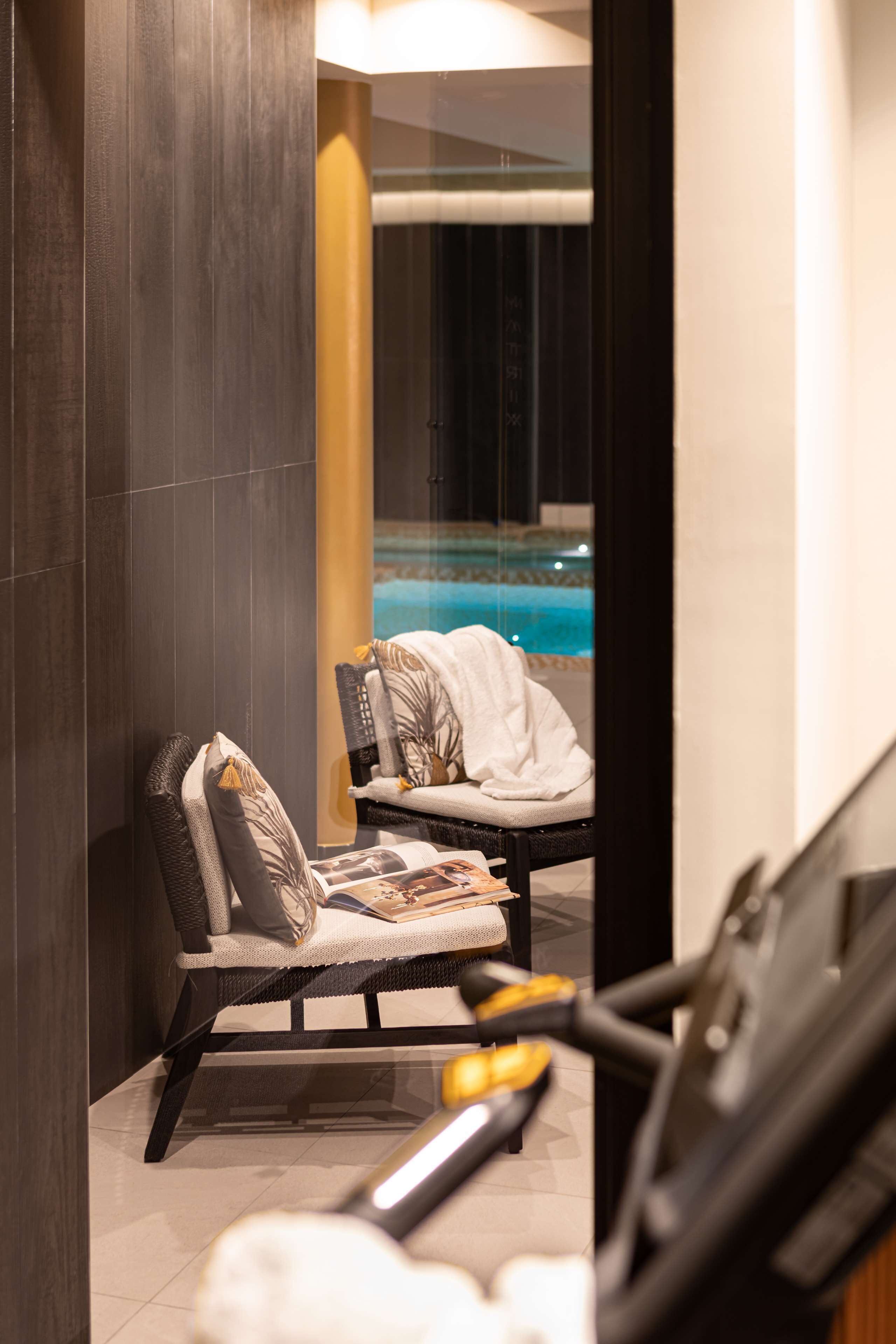 A modern room with a treadmill, a chair with a magazine, and a view of an indoor pool.