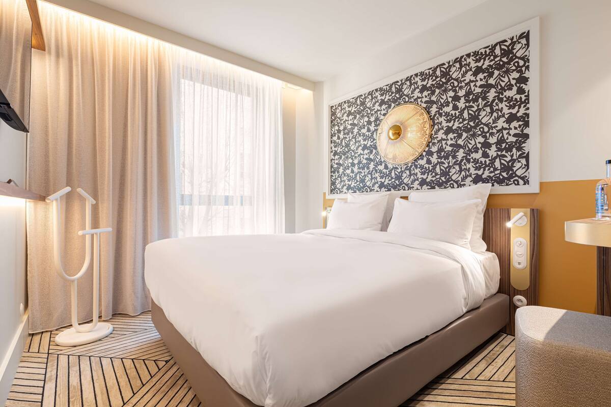 A hotel room with a large bed, a bedside table, a wall-mounted artwork, a floor lamp, and a window with curtains.