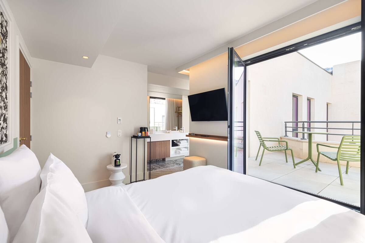 A hotel room with a bed, a small desk with a mirror, a TV mounted on the wall, and a balcony with chairs.