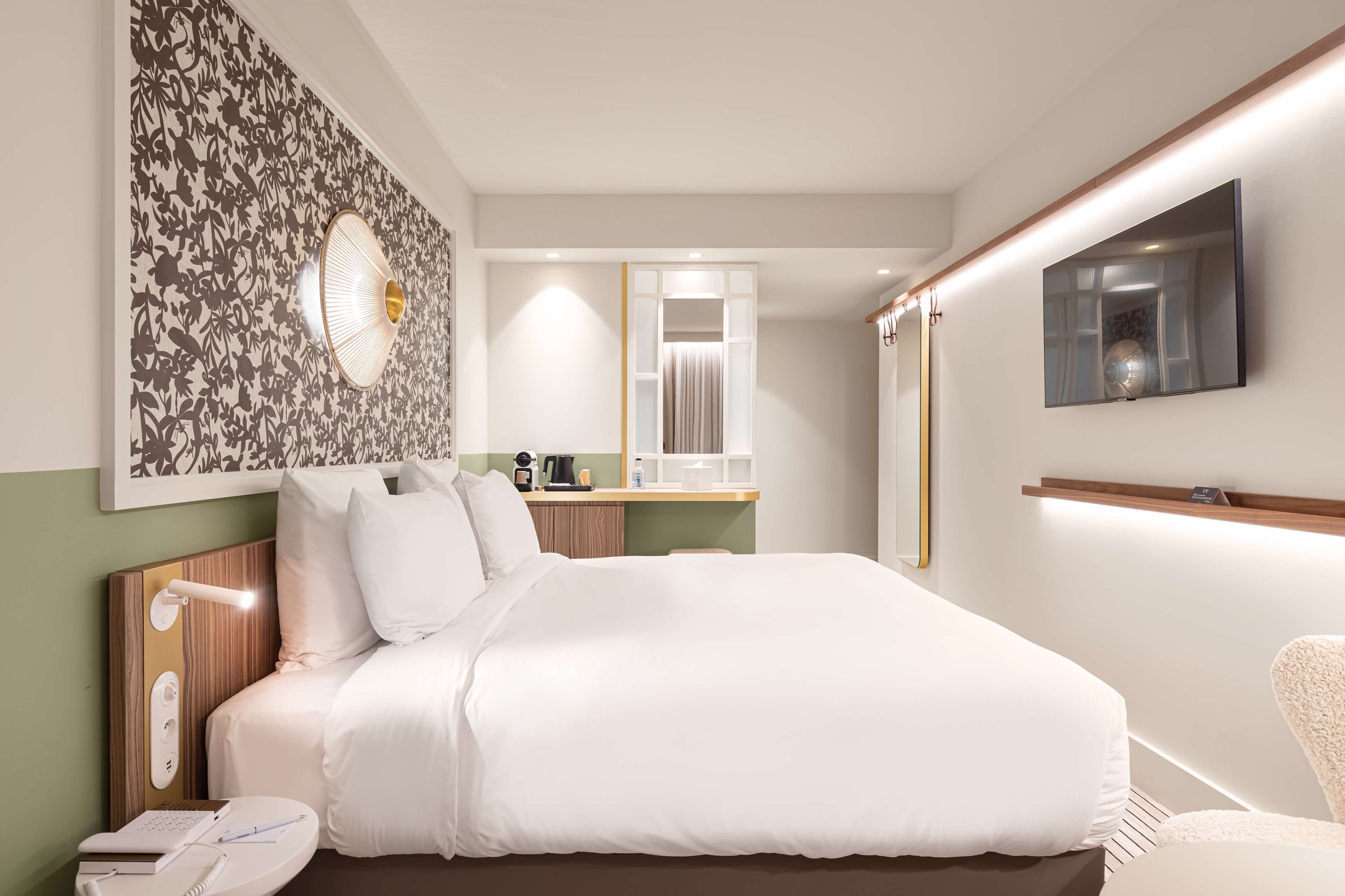 A modern hotel room with a large bed, a bedside table with a lamp, a vanity with a mirror, and a flat-screen TV mounted on the wall.