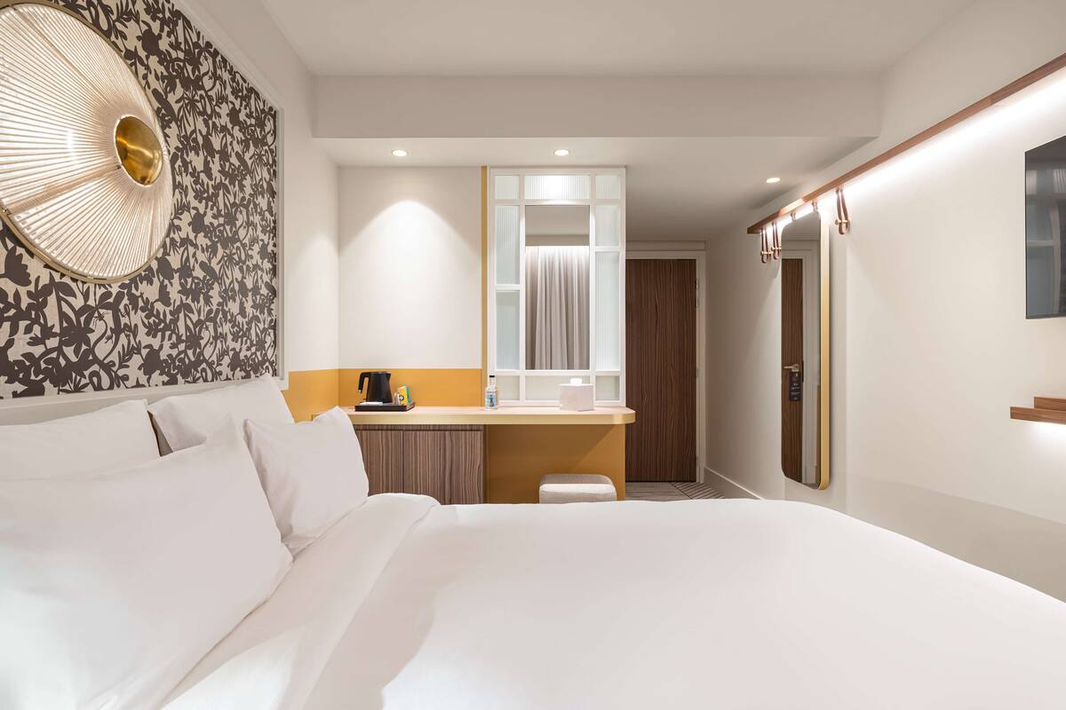 A hotel room with a large bed, a small desk with a mirror, a wall-mounted lamp, and a door with a wooden panel.