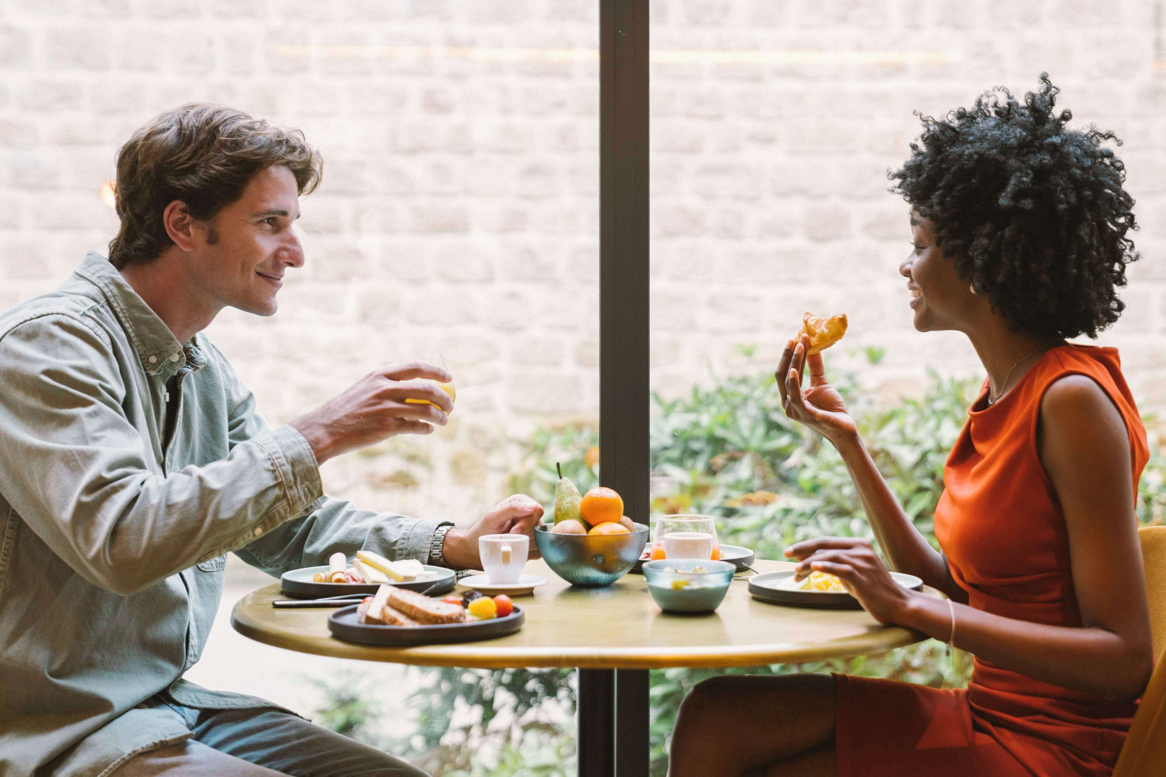 A couple dining at a table with a bowl of fruit, plates of food, and a cup.