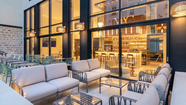 An outdoor seating area with sofas and tables, a bar with bottles and glasses, and a view of the interior through large windows. - Hotel Yac Paris Clichy, A Member Of Radisson Individuals (Clichy)
