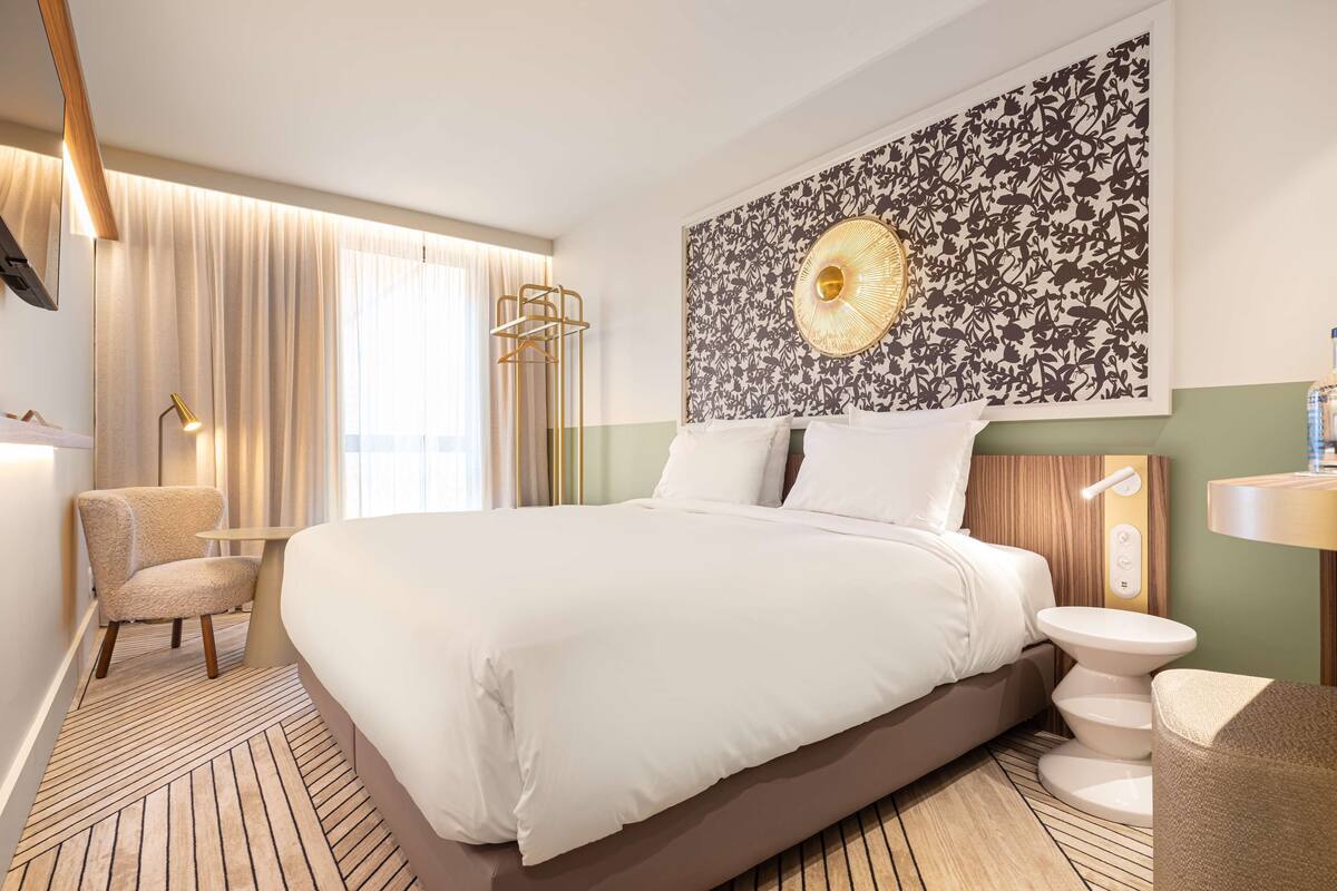 A modern hotel room with a large bed, a desk, and a chair. There is a window with curtains, a wall art piece, and a small table with a lamp.