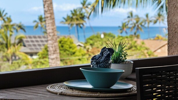 Ein Holztisch mit einer blauen Schüssel und einem Topfpflanzen auf einem Balkon mit Blick auf eine tropische Landschaft, Palmen und das Meer.