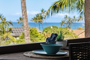 Een houten tafel met een blauwe kom en een potplant op een balkon met uitzicht op een tropisch landschap met palmbomen en de oceaan.