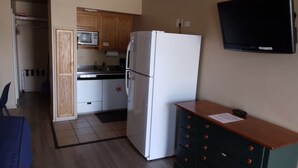 A compact kitchen with a refrigerator, microwave, and wooden cabinets. A flat-screen TV is mounted on the wall.