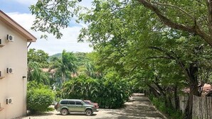 A residential street with a parked SUV, a house with air conditioning units, and lush greenery. - Tropical Hideaway 2+2 Condo in Playa Coco Guanacaste (Ocotal, Sardinal, Carrillo)