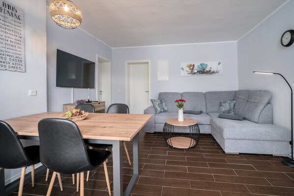 A modern living room with a grey sofa, a wooden dining table with chairs, a small side table with a vase, and a flat-screen TV mounted on the wall.