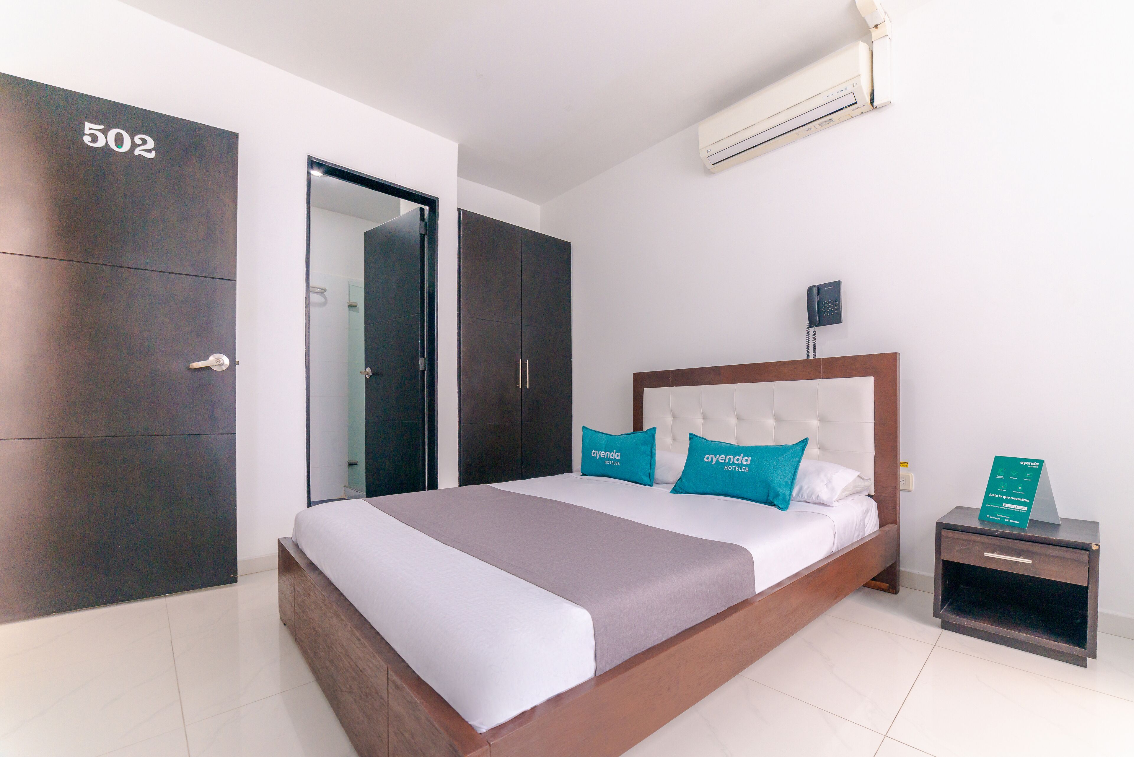 Habitación de hotel con una cama grande, un armario, una mesita de noche con un teléfono y un cartel con el nombre del hotel.