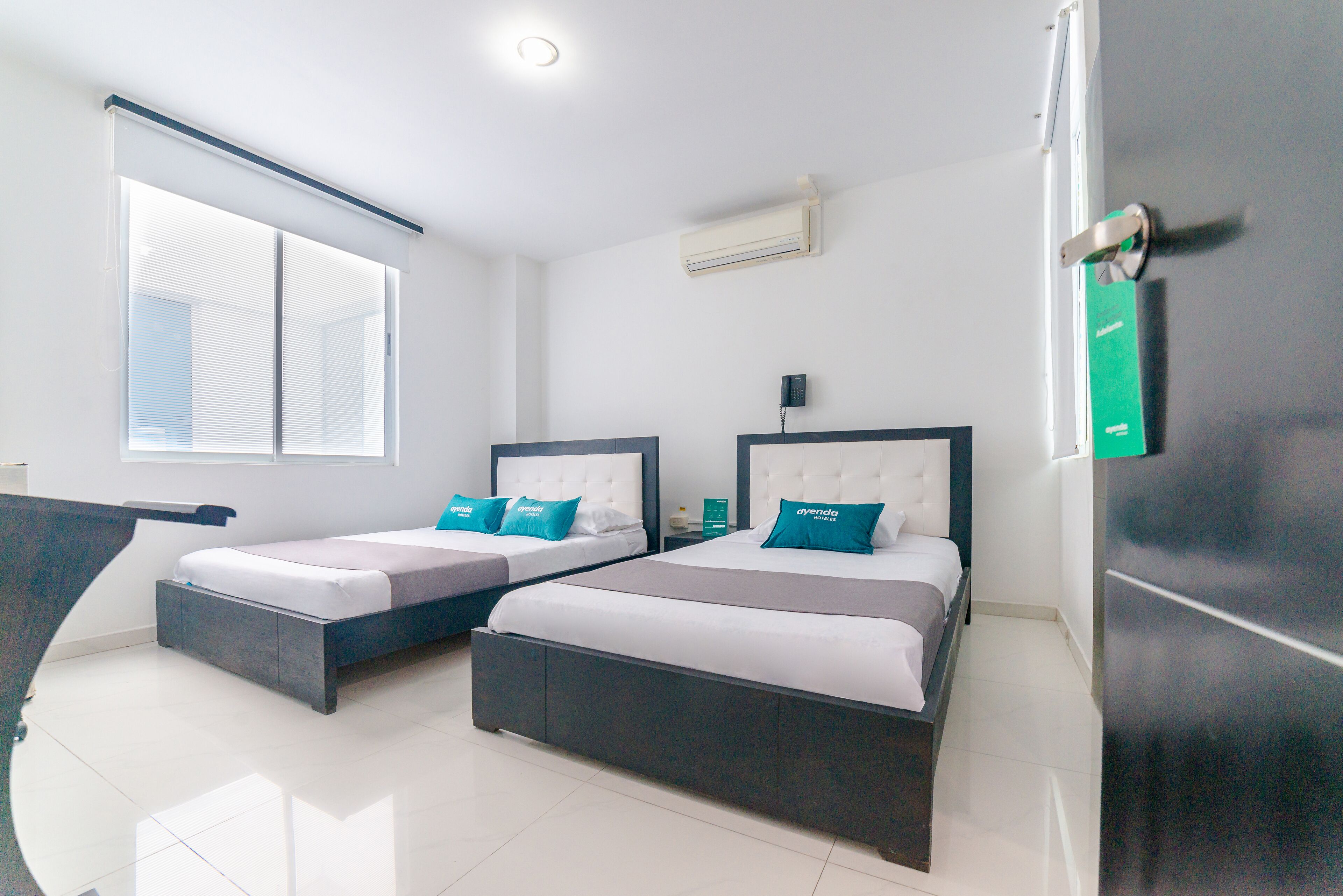 A hotel room with two beds, a window, an air conditioning unit, and a door with a green sign.