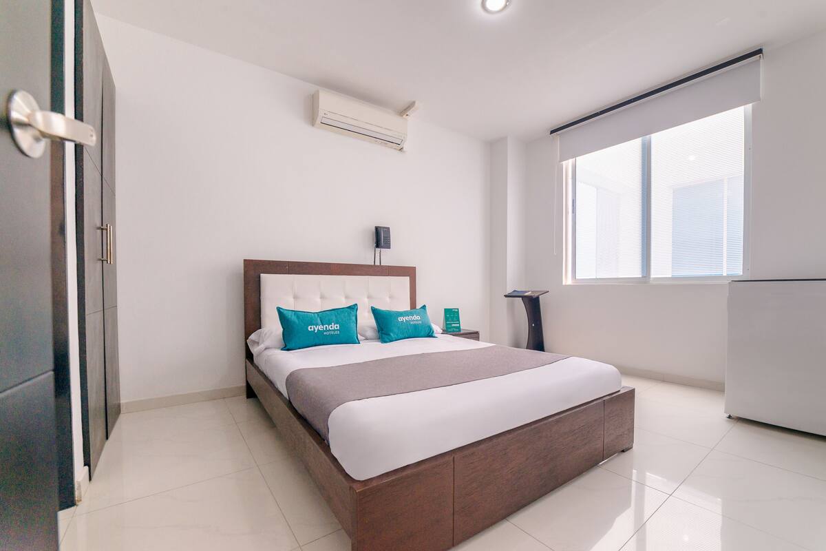 Quarto de hotel moderno com uma cama grande, uma mesa de cabeceira, um aparelho de ar condicionado e uma janela com persianas.