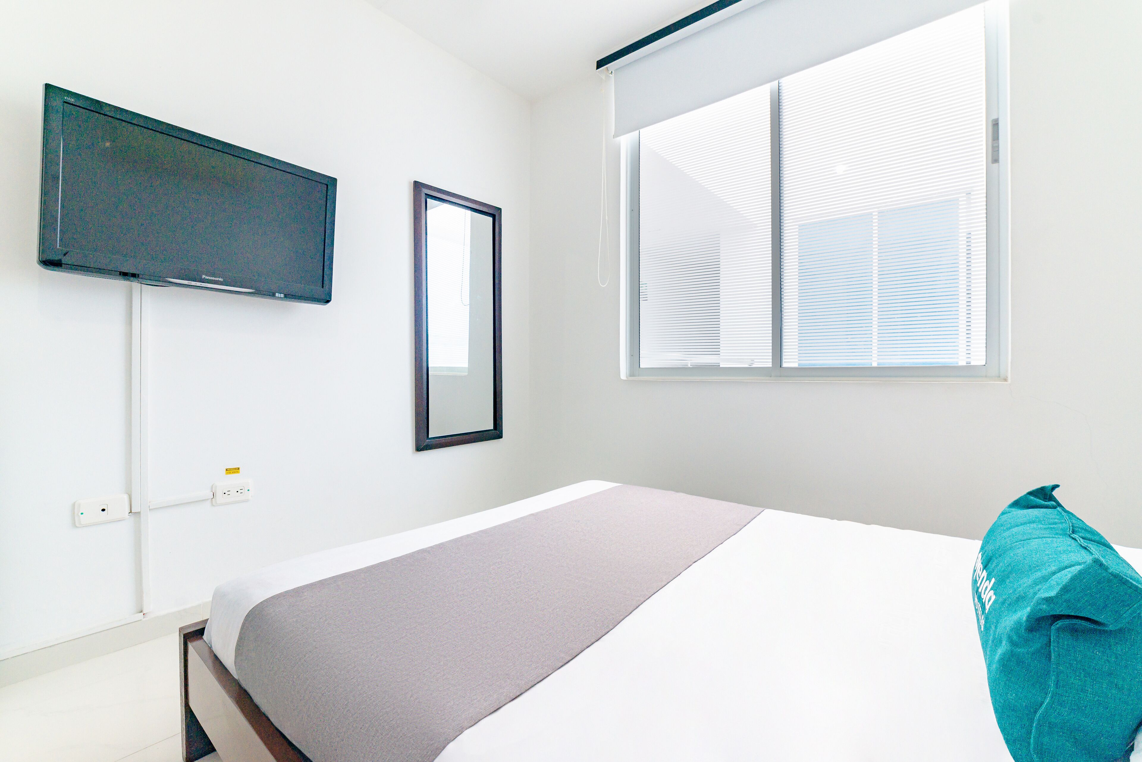 A bedroom with a bed, a TV mounted on the wall, a window with blinds, and a power outlet.