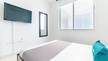 A bedroom with a bed, a TV mounted on the wall, a window with blinds, and a power outlet.