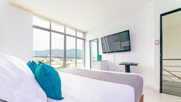 Una habitación de hotel con una cama, un televisor montado en la pared y una ventana con vistas a edificios y colinas.