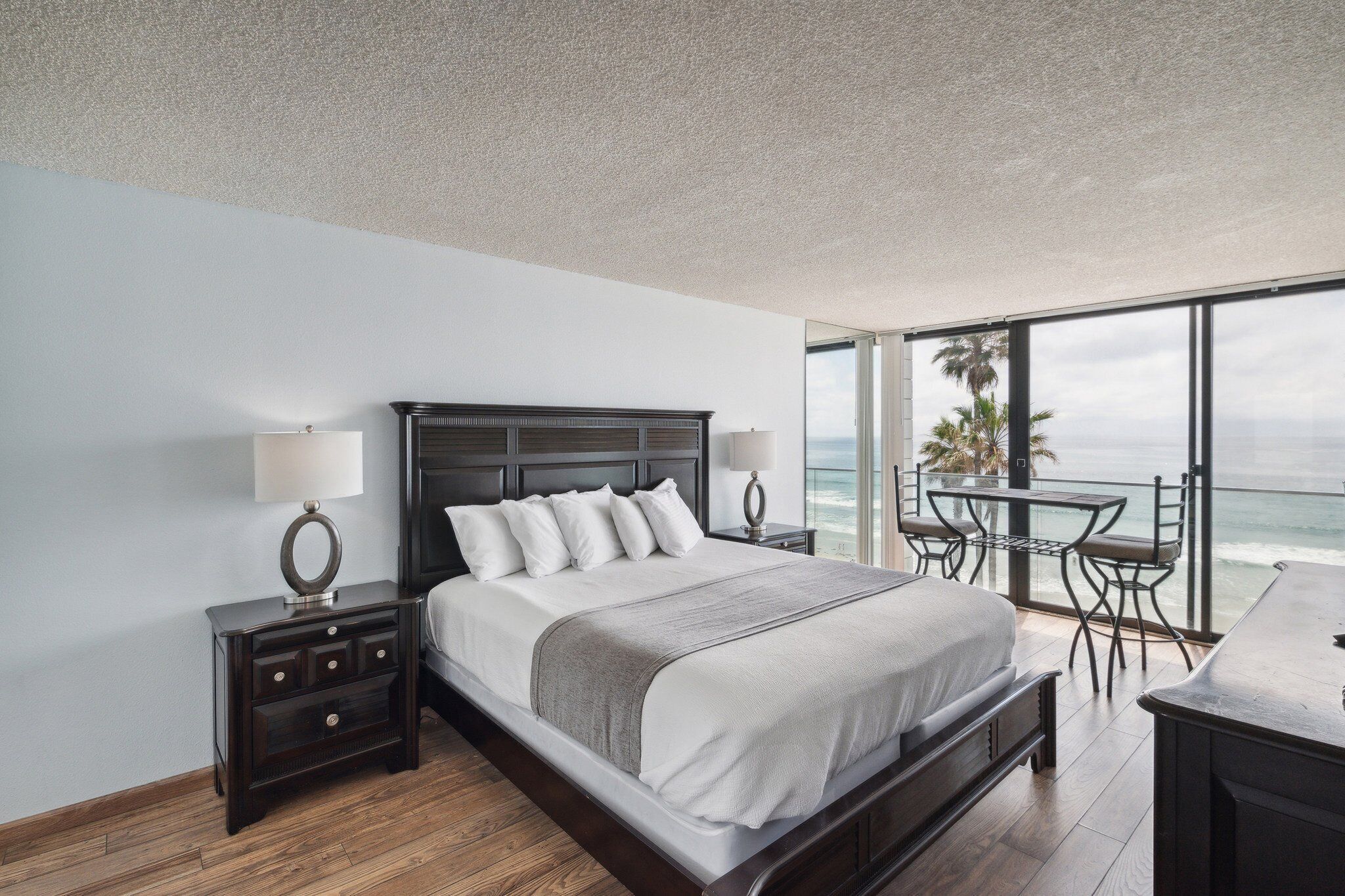 A modern bedroom with a large bed, bedside tables, a balcony with a table and chairs, and a view of the ocean.