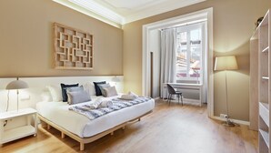 Un dormitorio moderno con una cama grande, mesitas de noche, un escritorio y vistas de la ciudad a través de una ventana.