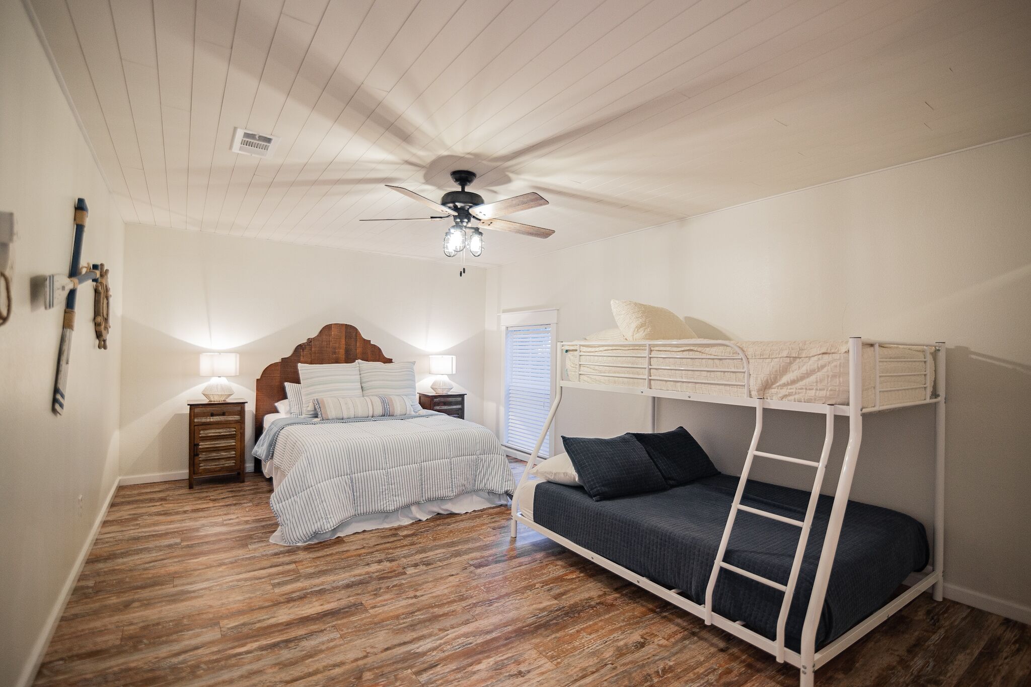 A bedroom with a bunk bed, a single bed, a wooden floor, a ceiling fan, and a nightstand with a lamp.