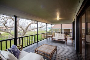 A covered porch with wicker furniture, a view of a tree-lined yard, and a building in the background.