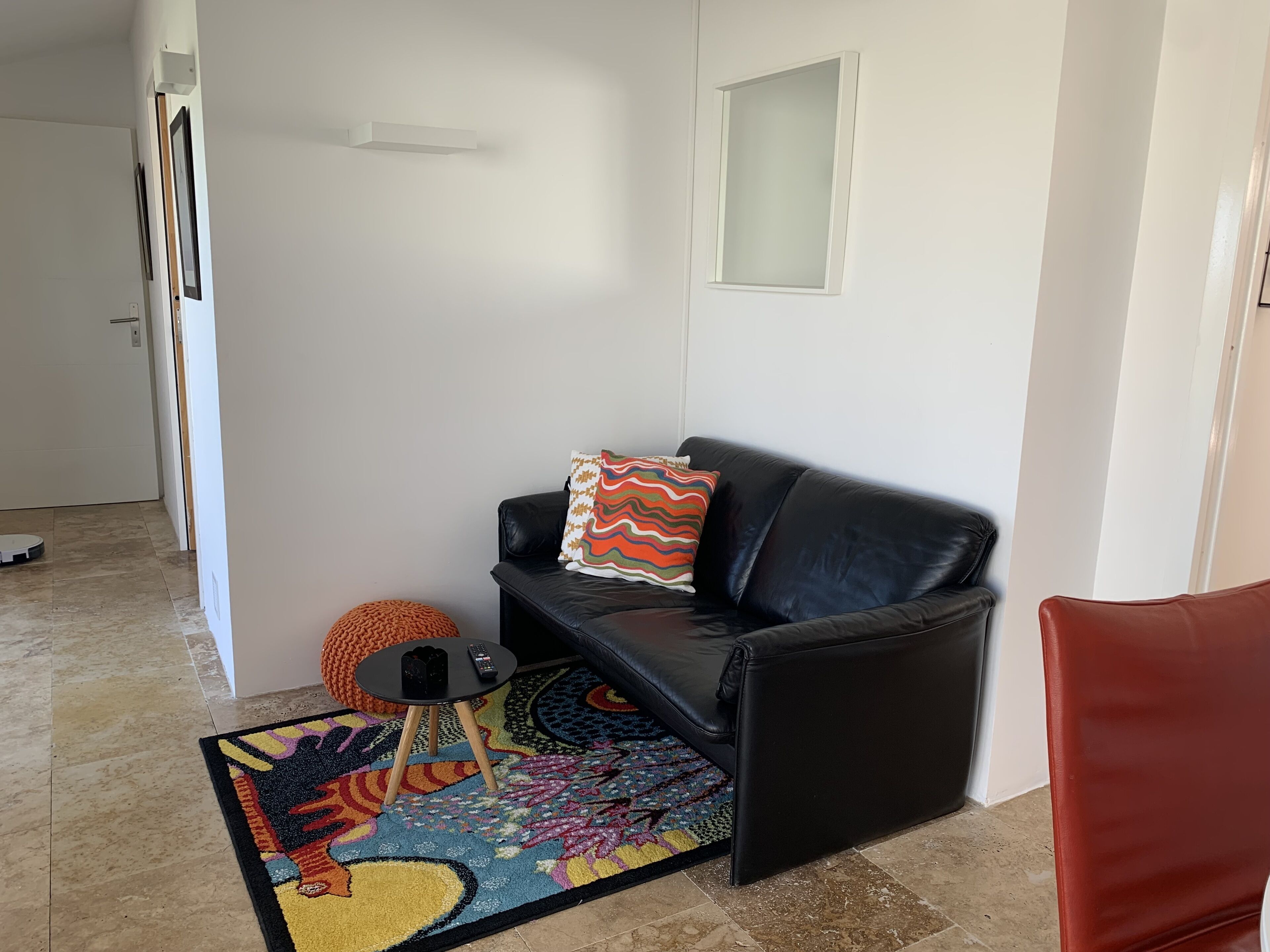 A living room with a black leather sofa, a colorful rug, a small round ottoman, and a remote control on a small table.