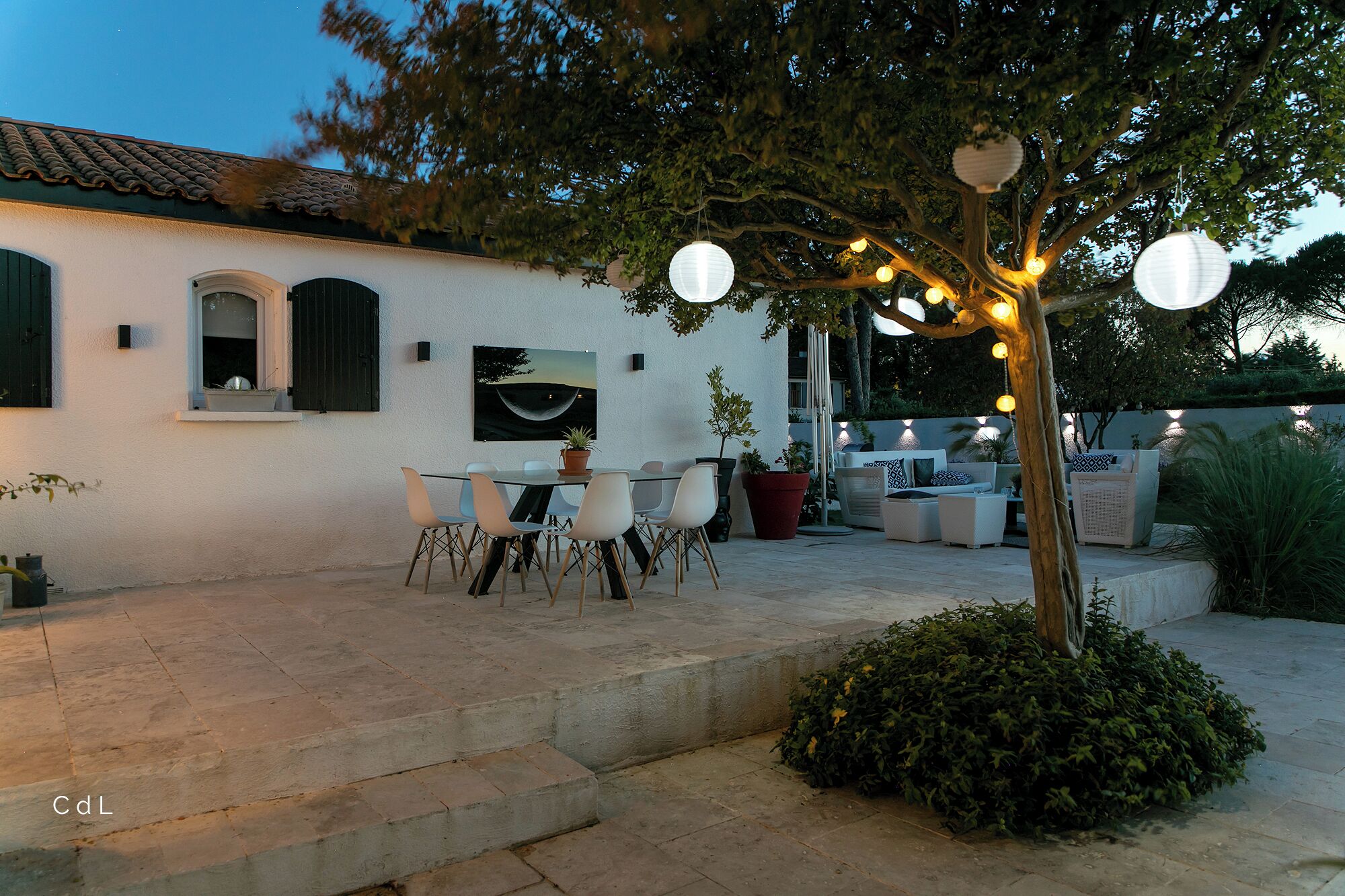 Un espace patio aménagé avec une table et des chaises, un pot de fleurs et un arbre décoré de guirlandes lumineuses.