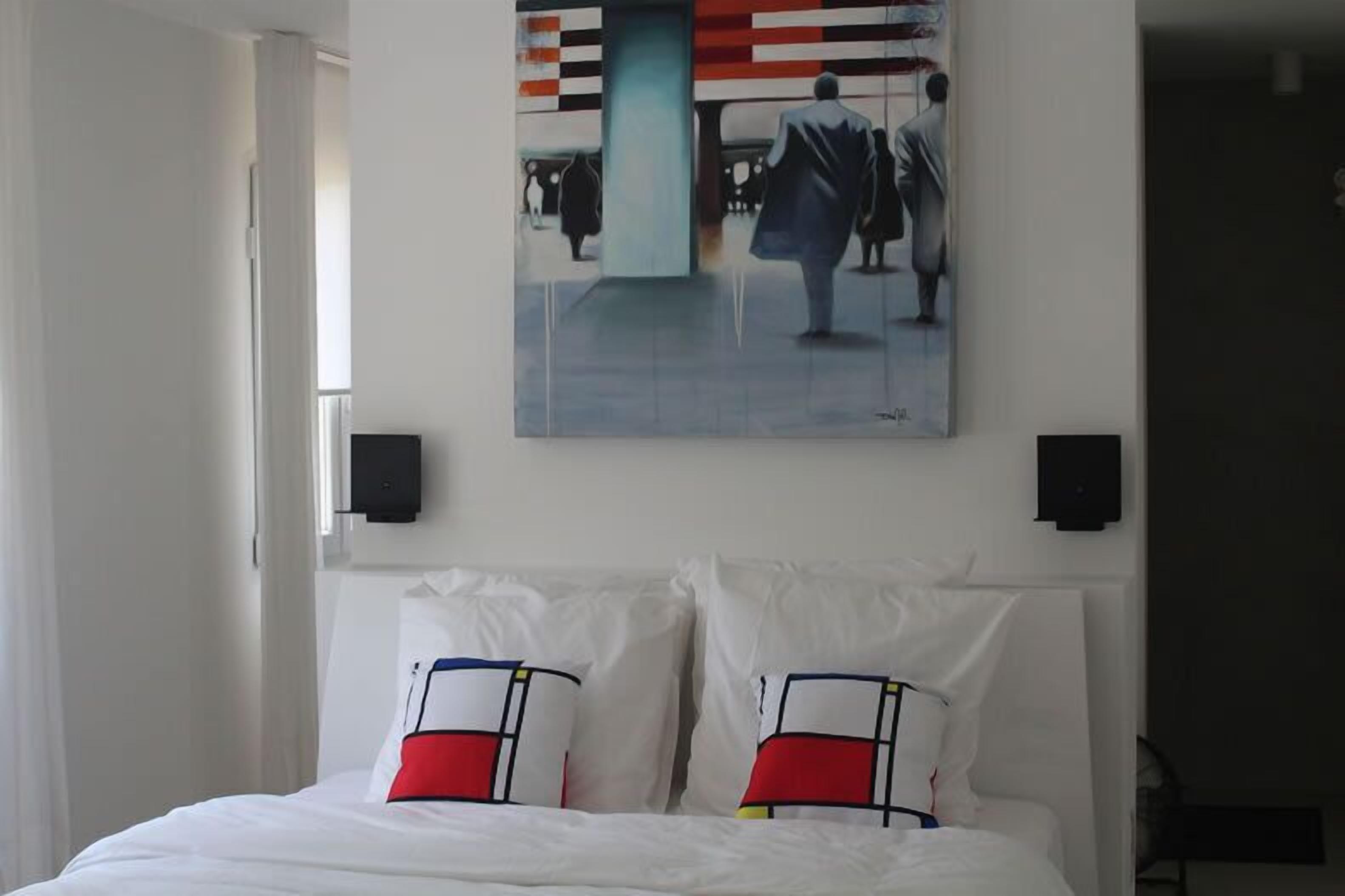 A hotel room with a bed, white pillows, a painting on the wall, and a window.