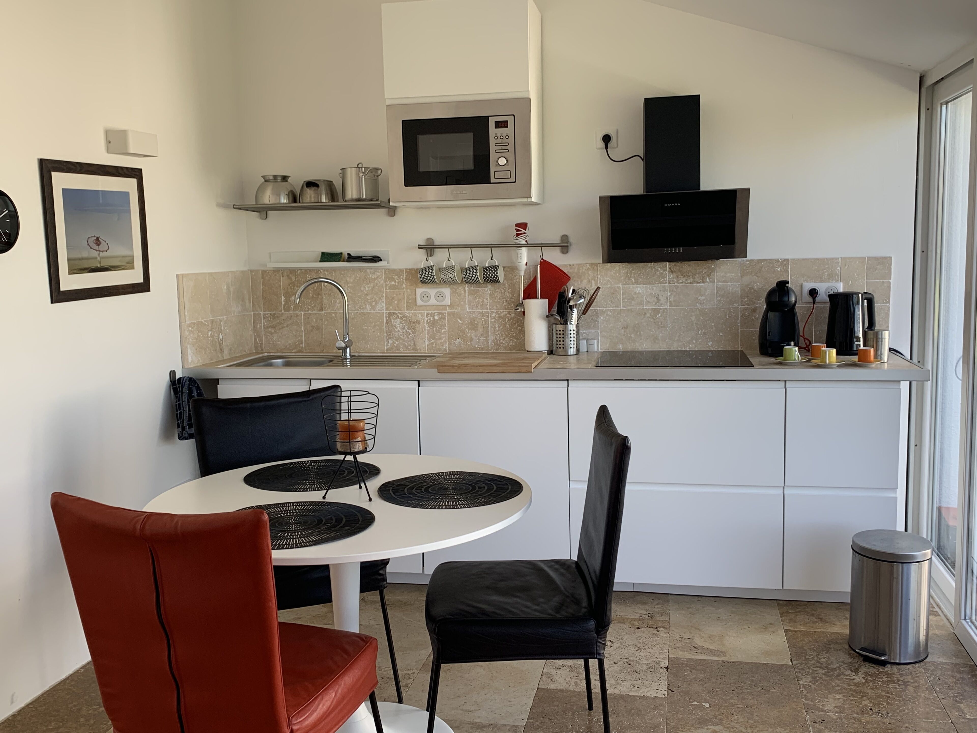 Cocina compacta con microondas, fregadero y cafetera. Hay una mesa de comedor con sillas y un pequeño cubo de basura.