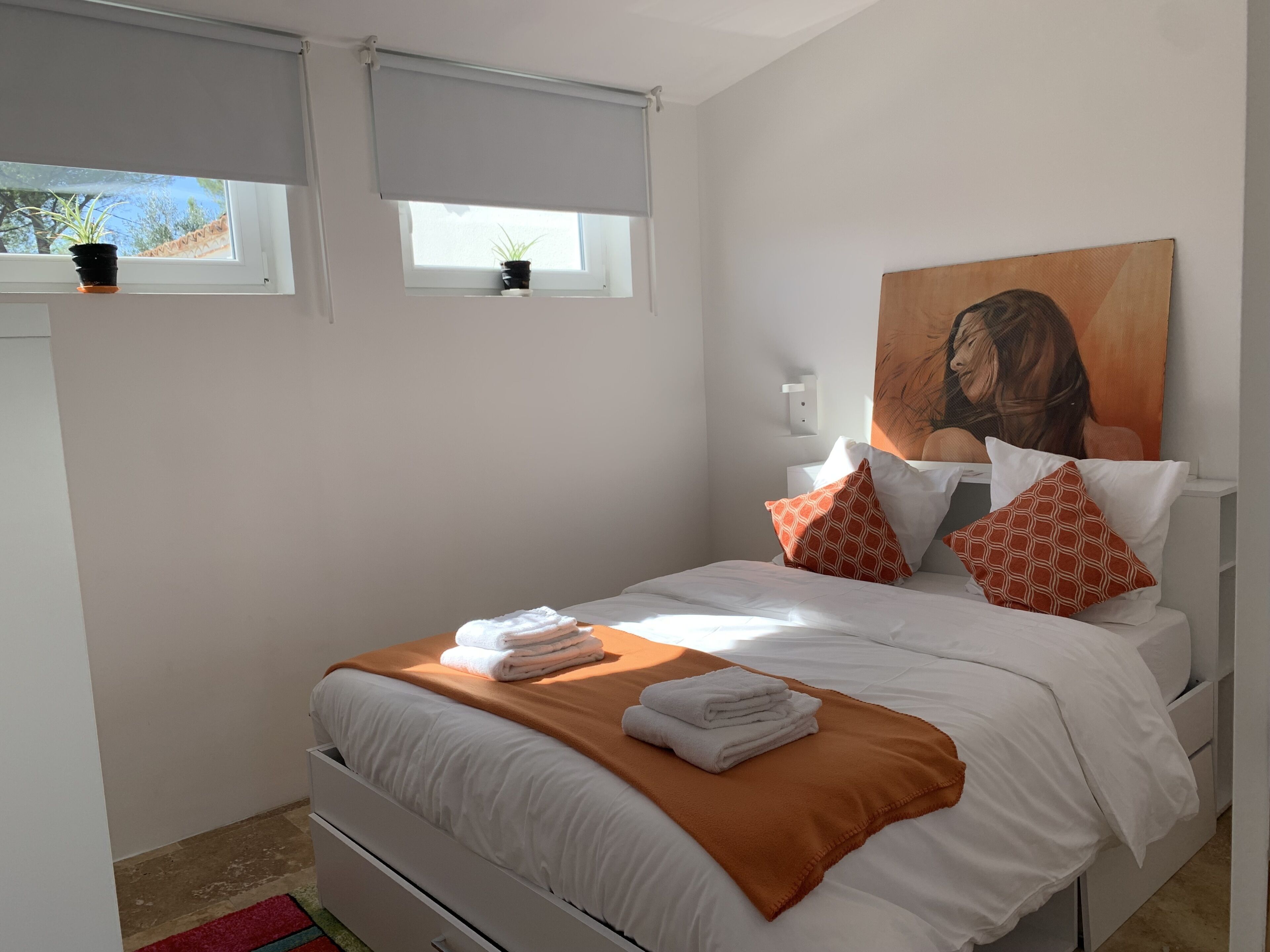 Una cama bien hecha con ropa de cama blanca y naranja, un cuadro en la pared y una ventana con persianas.