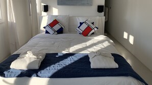 A neatly made bed with a blue blanket and two pillows, a white wall with a painting, and a window with curtains. - Croissant de Lune (Bagard)