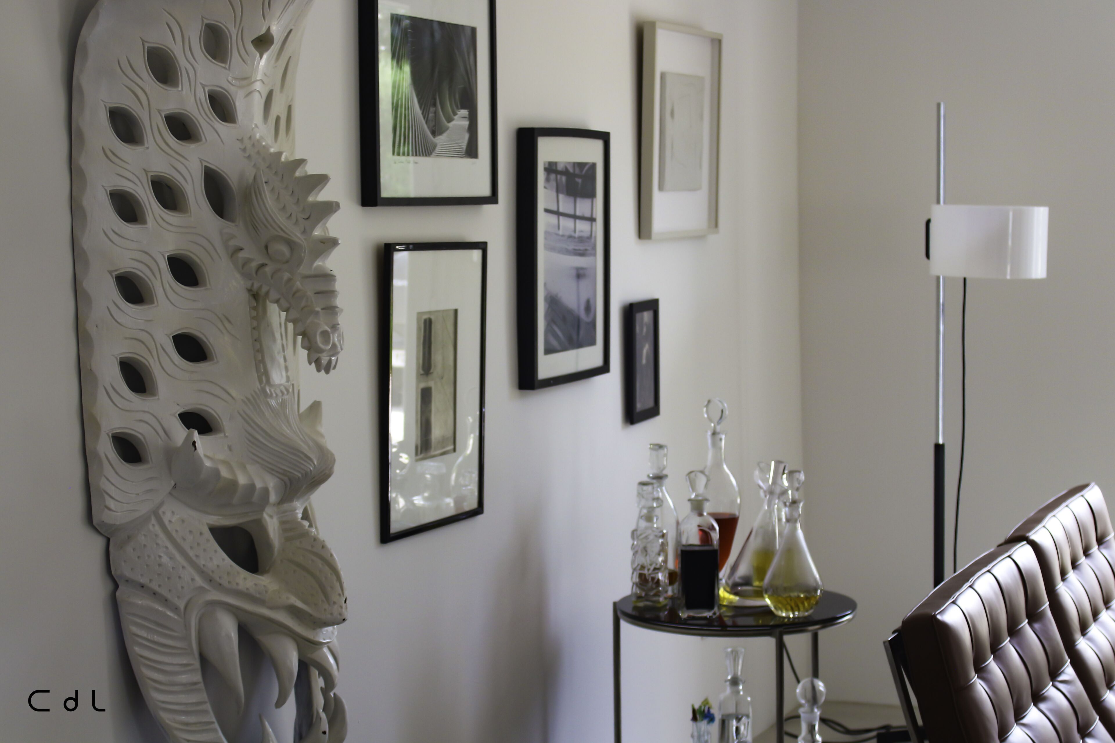 Een kamer met een witte decoratieve wandversiering, ingelijste foto's, een klein tafeltje met glazen en een bruine leren fauteuil.