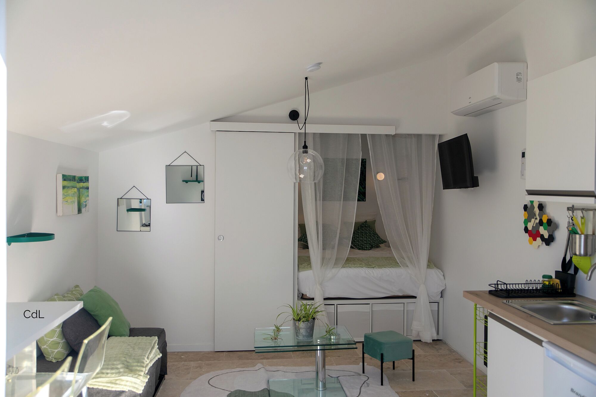 A compact living space with a glass dining table, a bed with white curtains, a kitchenette, and a small seating area with a chair and cushions.
