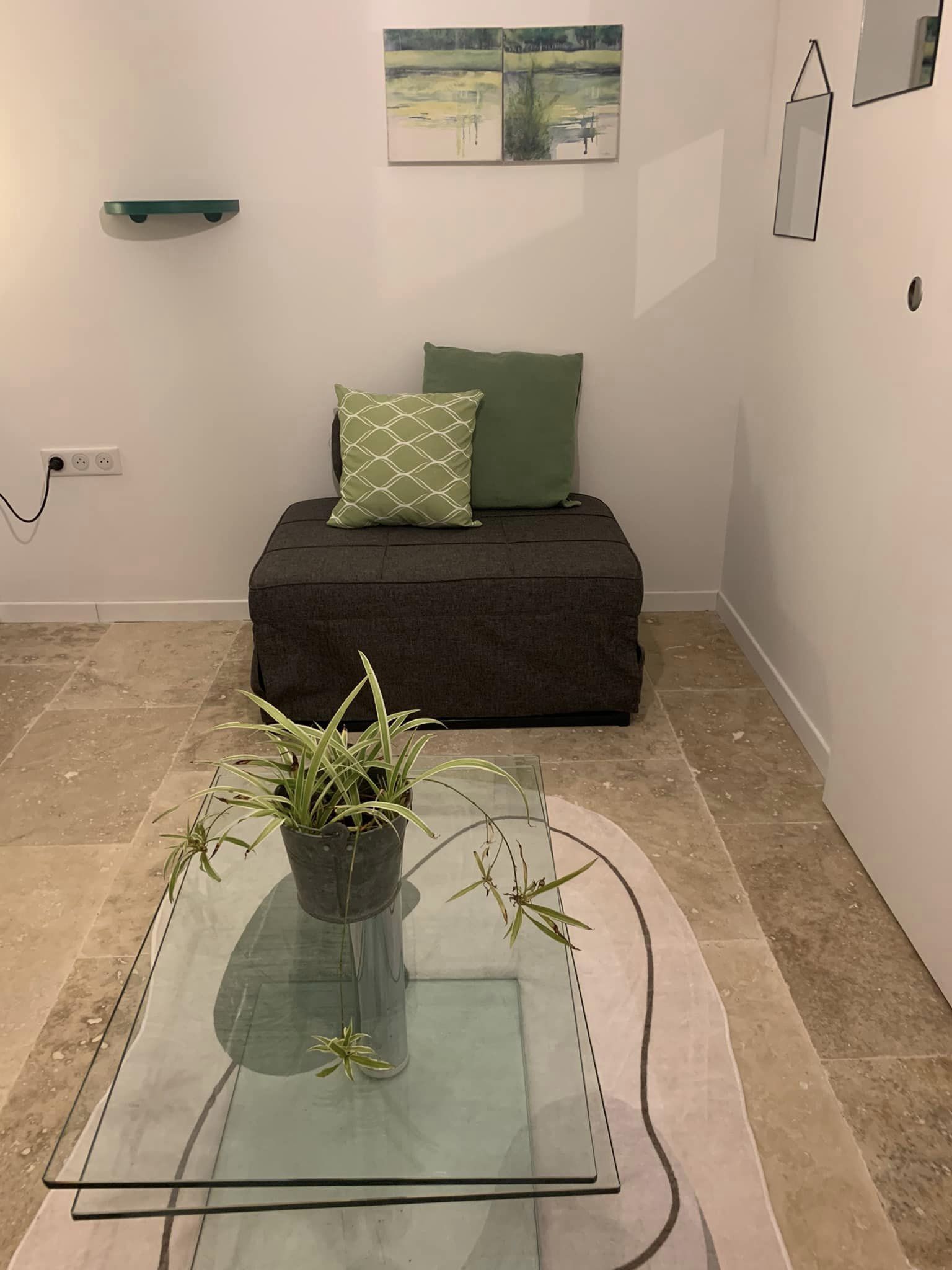 A room with a glass-top table, a dark sofa with green cushions, a wall-mounted shelf, and a painting.