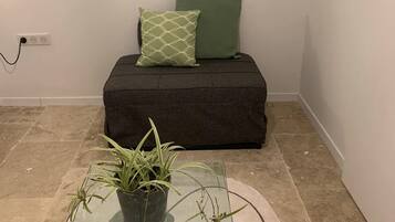 Una habitación con una mesa de vidrio, un sofá oscuro con cojines verdes, un estante montado en la pared y un cuadro.