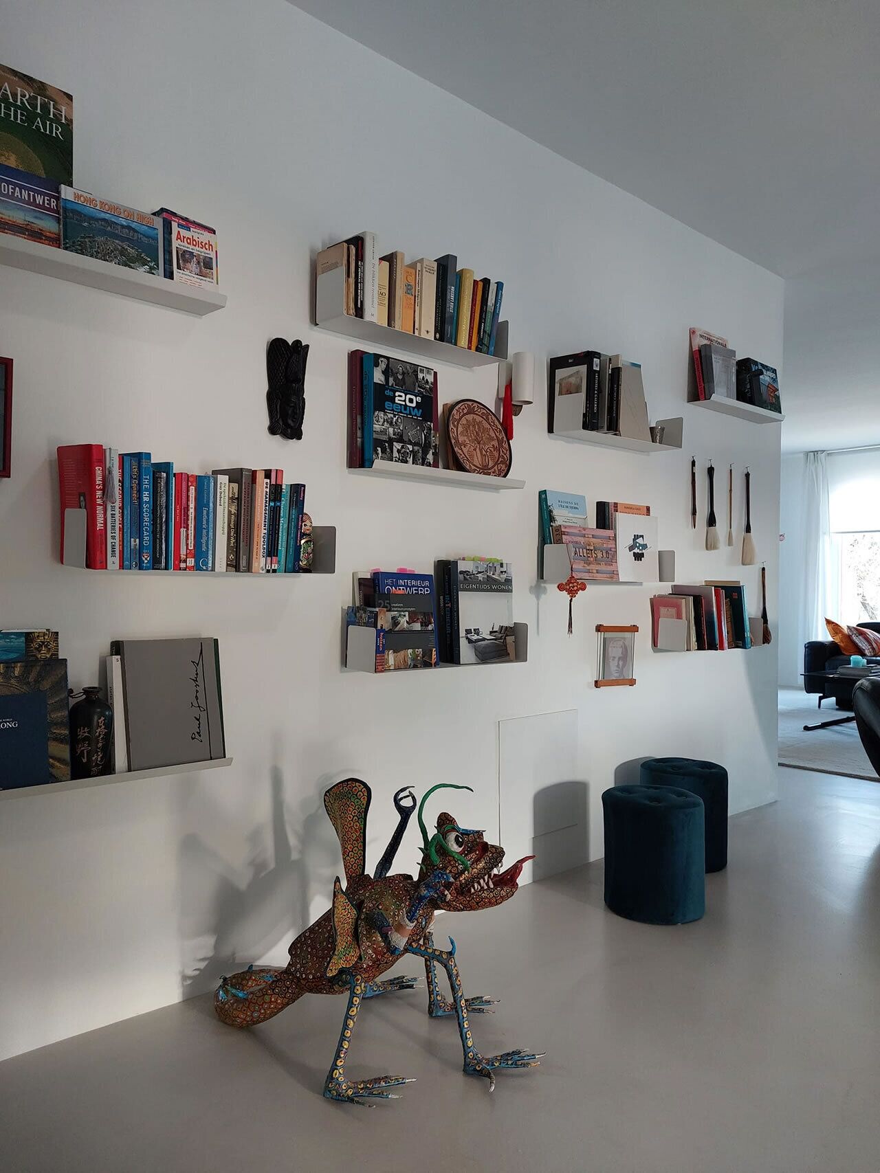 A modern living room with a white wall adorned with bookshelves containing various books and decorative items. A unique sculpture is placed on the floor, and there are two blue ottomans.