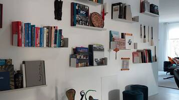 A modern living room with a white wall adorned with bookshelves containing various books and decorative items. A unique sculpture is placed on the floor, and there are two blue ottomans.