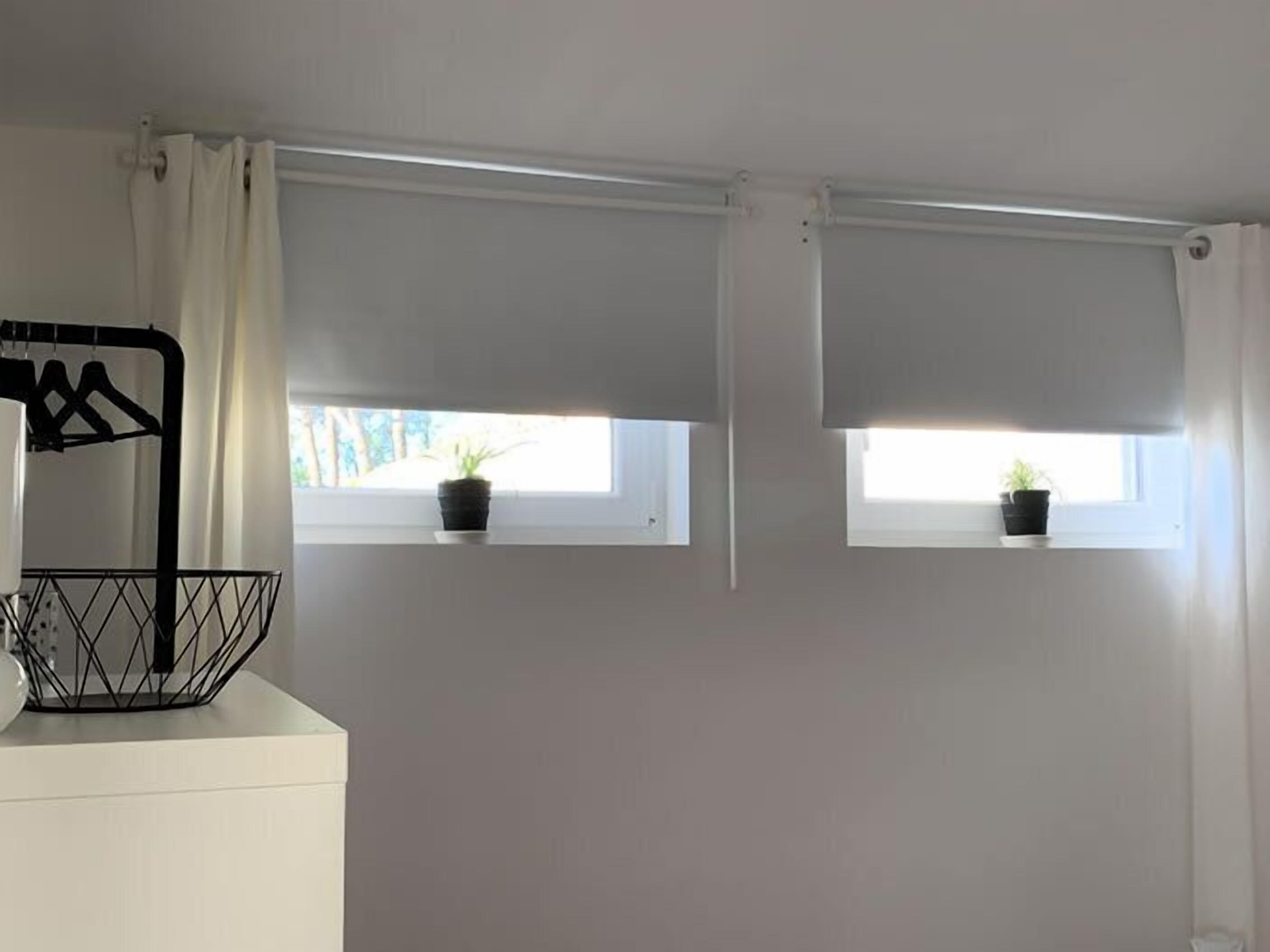Een kamer met twee ramen, elk voorzien van een plant op de vensterbank en een wit gordijn. Er staat een zwarte metalen mand op een witte sokkel.