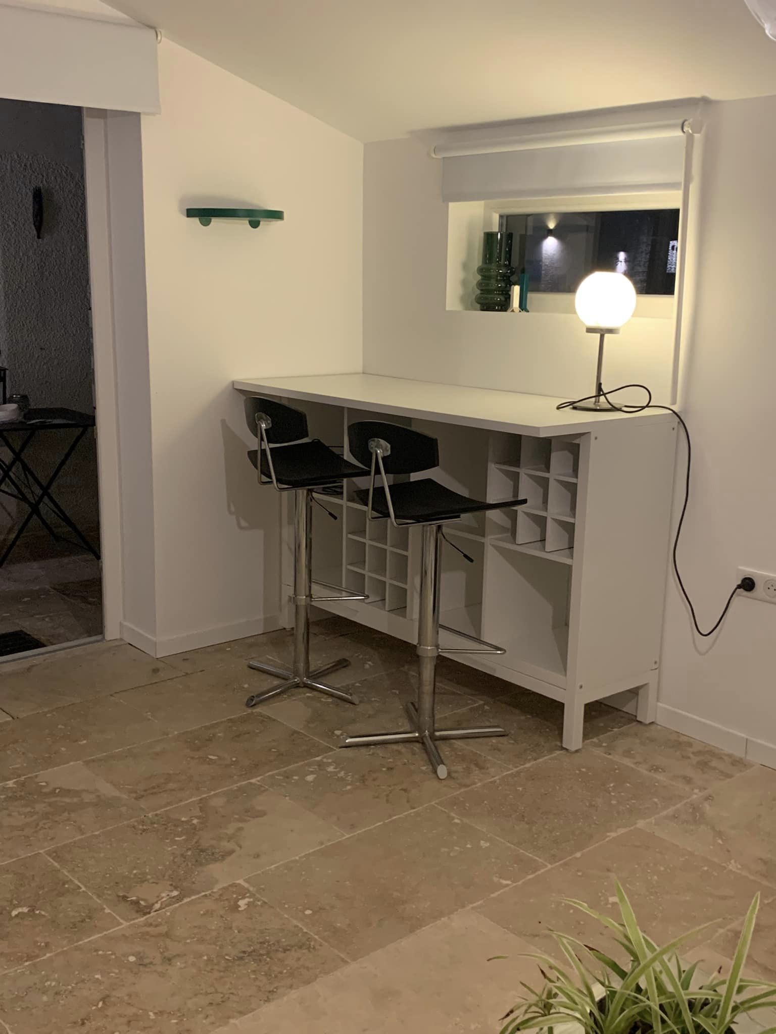 A small kitchenette with a white countertop, two black bar stools, a mirror, and a wall-mounted light.