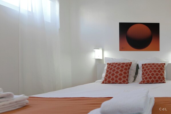 Habitación de hotel con cama, ropa de cama blanca, almohadas rojas estampadas, una obra de arte colgada en la pared y una lámpara de noche.