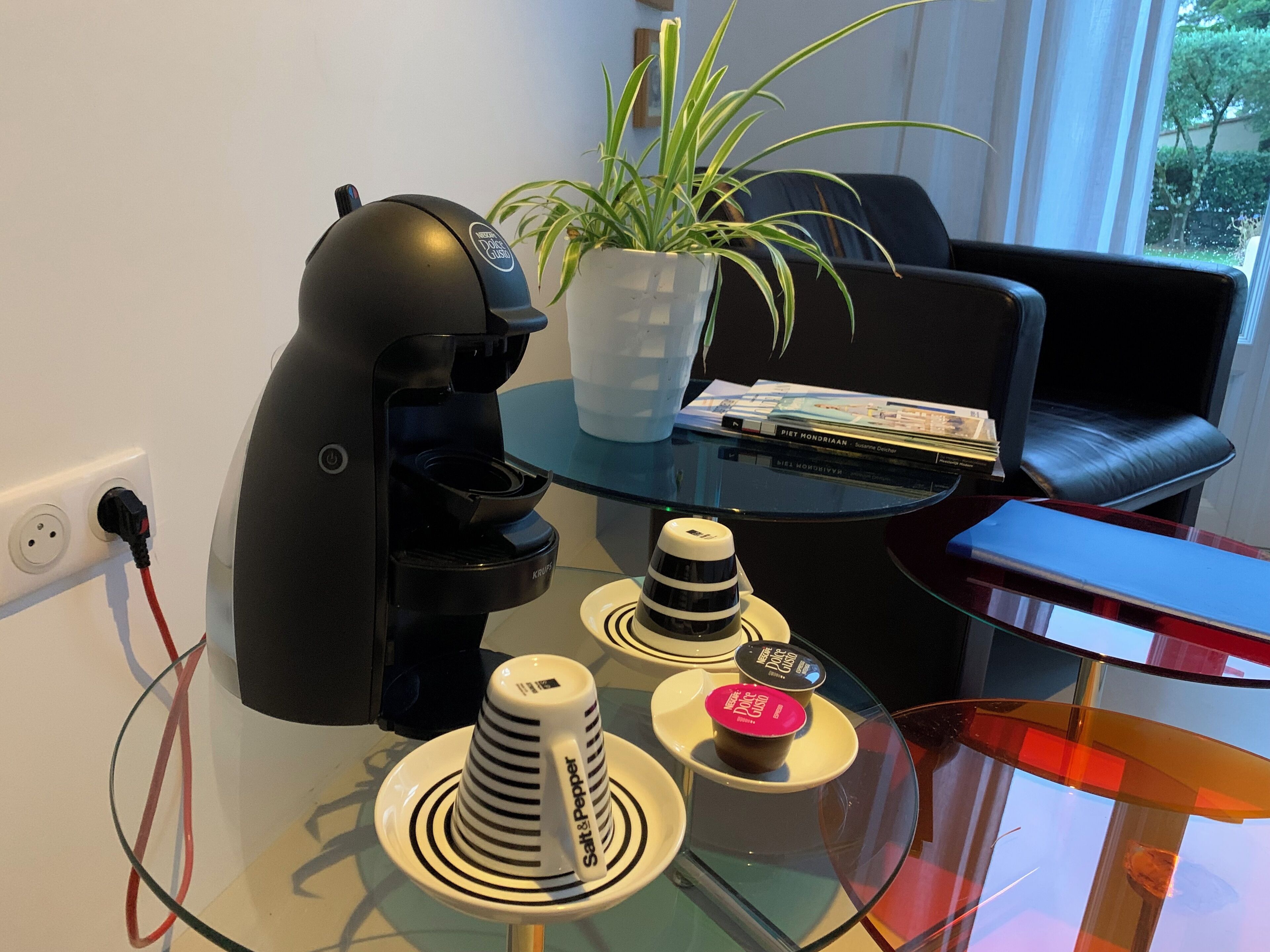 Una habitación moderna con una cafetera negra, una mesa de vidrio con tazas apiladas, una planta en maceta y un sillón de cuero.