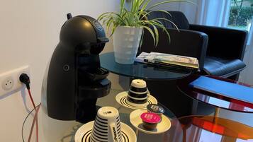Una habitación moderna con una cafetera negra, una mesa de vidrio con tazas apiladas, una planta en maceta y un sillón de cuero.