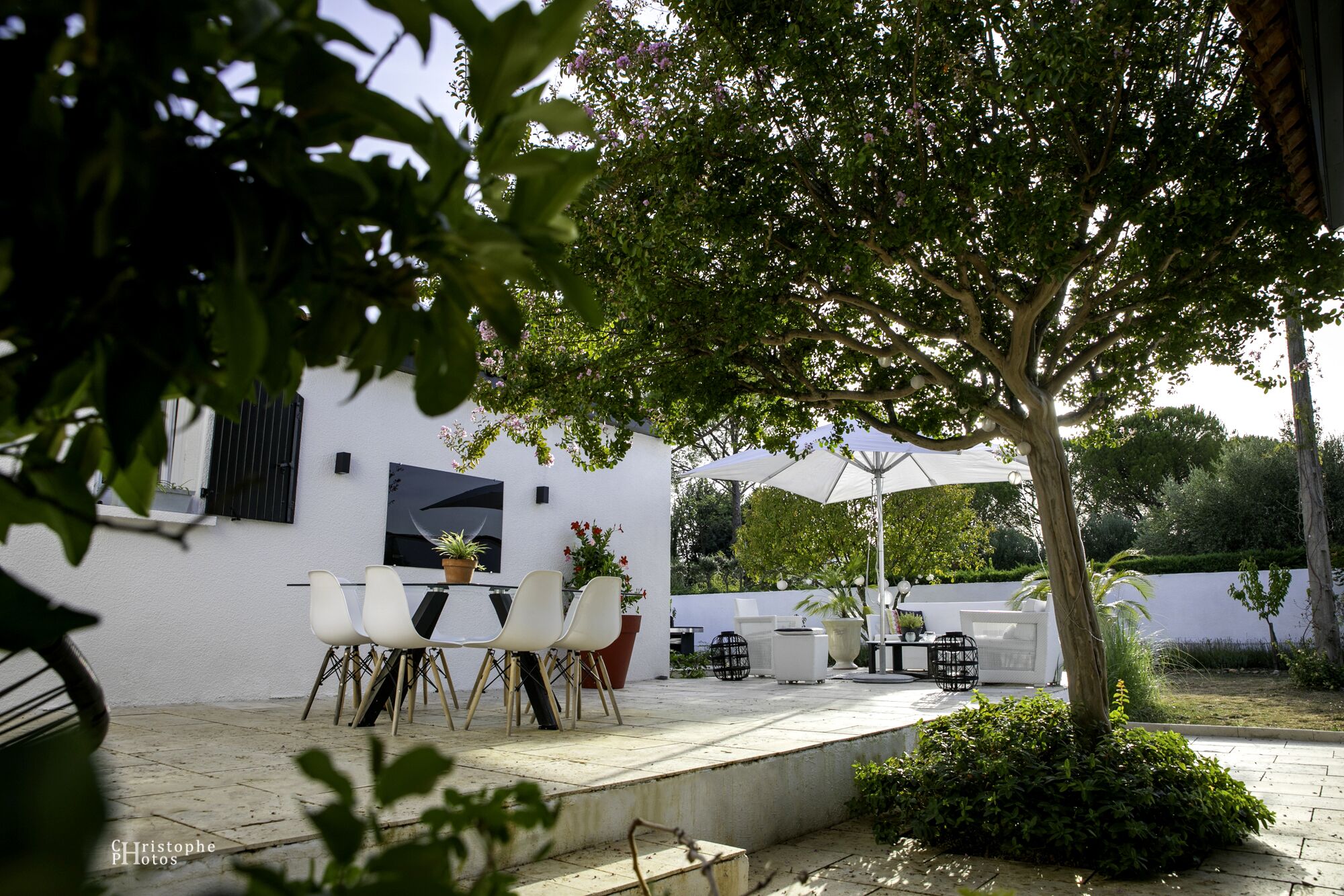An outdoor patio with white chairs and tables, surrounded by greenery and a white building with a black-framed window.