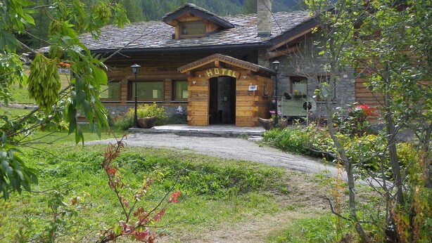 A rustic wooden hotel with a stone chimney, a welcoming entrance, and a view of a forested mountain in the background. - Hotel Les Granges (La Thuile)