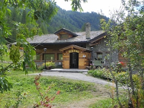A rustic wooden hotel with a stone chimney, a welcoming entrance, and a view of a forested mountain in the background. - Hotel Les Granges (La Thuile)