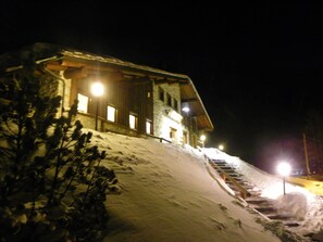 Une pente enneigée menant à un chalet en bois doté de nombreuses fenêtres et d’un escalier.