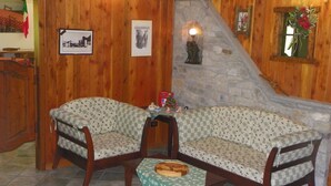 A cozy room with wooden walls, a stone fireplace, two patterned chairs, a small table with a bread basket, and a framed picture on the wall. - Hotel Les Granges (La Thuile)