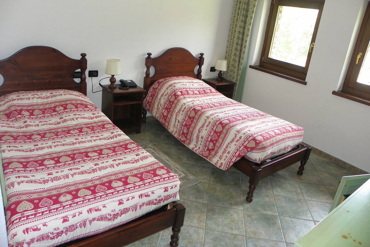 Una habitación con dos camas de madera, cada una con una colcha estampada. Hay mesitas de noche con lámparas y una pequeña mesita con un teléfono. Se ve una ventana con cortina en la pared.