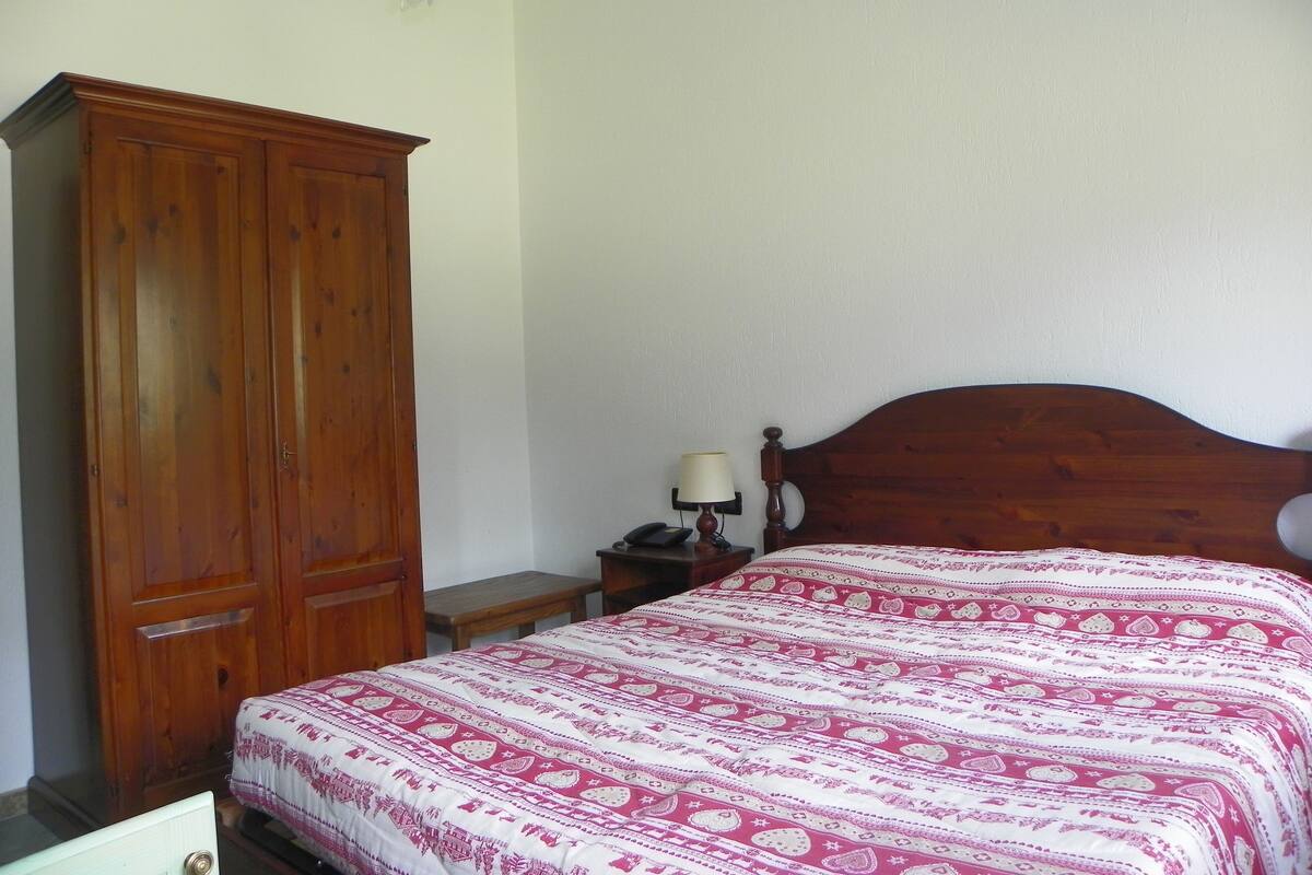 Un dormitorio con un armario de madera, una cama con colcha estampada, una mesita de noche con lámpara y un cabecero de madera.